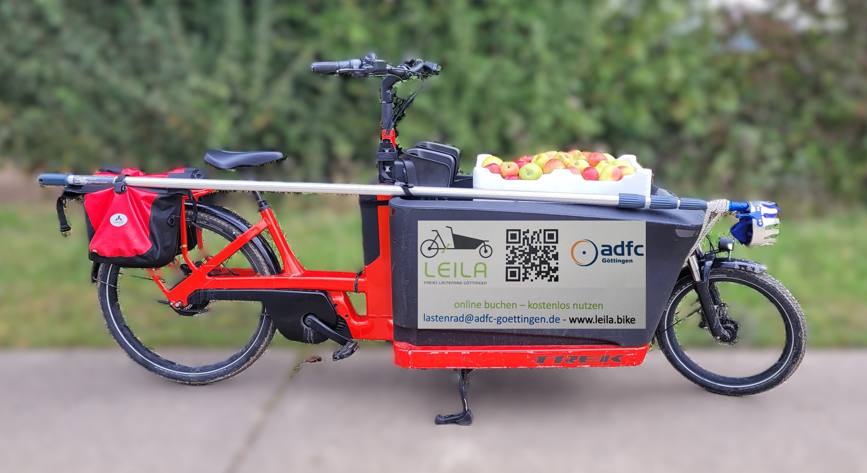 Das Bild zeigt ein rotes Lastenrad mit einer großen schwarzen Kiste mit lauter gepflückten Äpfeln vorne drauf.