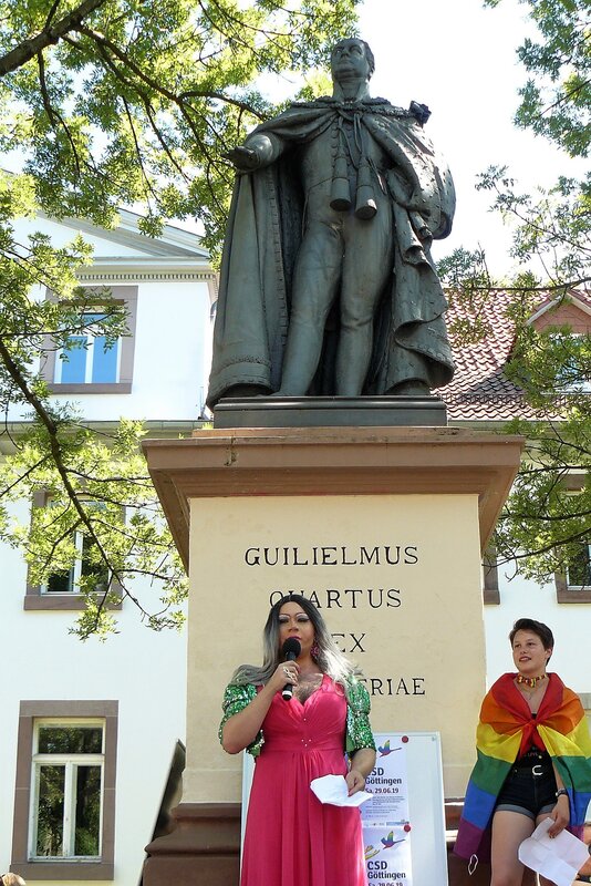 Drag Queen spricht in ein Mikrofon und steht vor einem Denkmal.