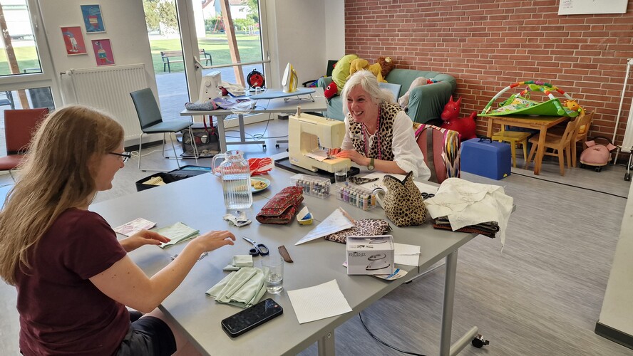 Zwei Frauen sitzen sich an einem Tisch gegenüber, nähen und lachen dabei.