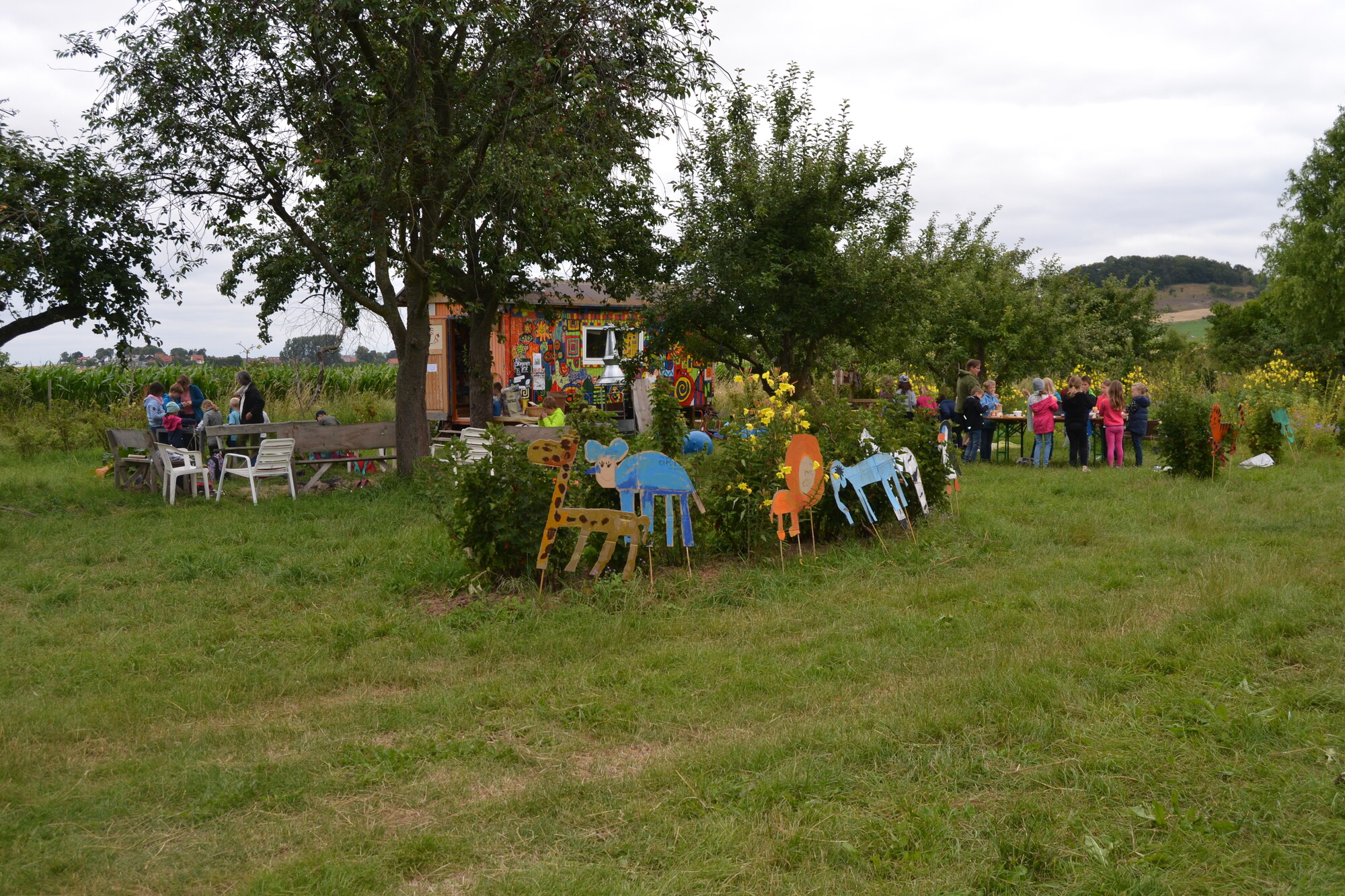 Ein Garten mit bunten Pappfiguren im Vordergrund, Büsche und Bäume, ein bunter Bauwagen und Kindergruppen an Tischen arbeitend.
