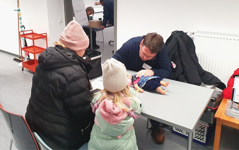 Ein Reparateur des Repair-Cafés hilf einer Mutter und ihrer Tochter bei der Reparatur einer Puppe.