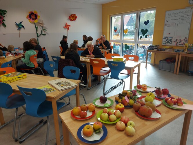 Spieltische mit Café-Besucher*innen. Im Vordergrund eine Ausstellung verschiedener Apfelsorten